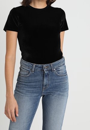 Mujer con una blusa ajustada de terciopelo negro de manga corta, metida dentro de unos vaqueros de talle alto azul con detalles descoloridos.