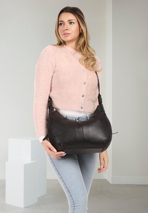 Vrouw in een roze, pluizige trui en lichte spijkerbroek die een grote zwarte leren schoudertas binnen vasthoudt.