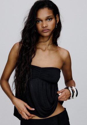 Mladá žena s dlhými vlnitými tmavými vlasmi má na sebe čierny top bez ramienok a veľké strieborné náramky, pózuje na jednoduchom svetlom pozadí.