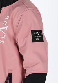 Lys pink jakke med en sort lap, der viser teksten "SLADE COUTURE". Lynlåslomme på siden, glat tekstur, moderne design.