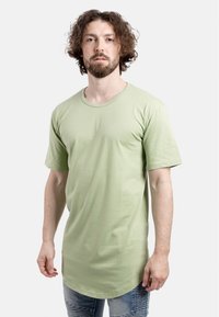 Camiseta de algodón verde claro con mangas cortas y escote redondo, que presenta un dobladillo ligeramente alargado y una textura suave y uniforme.