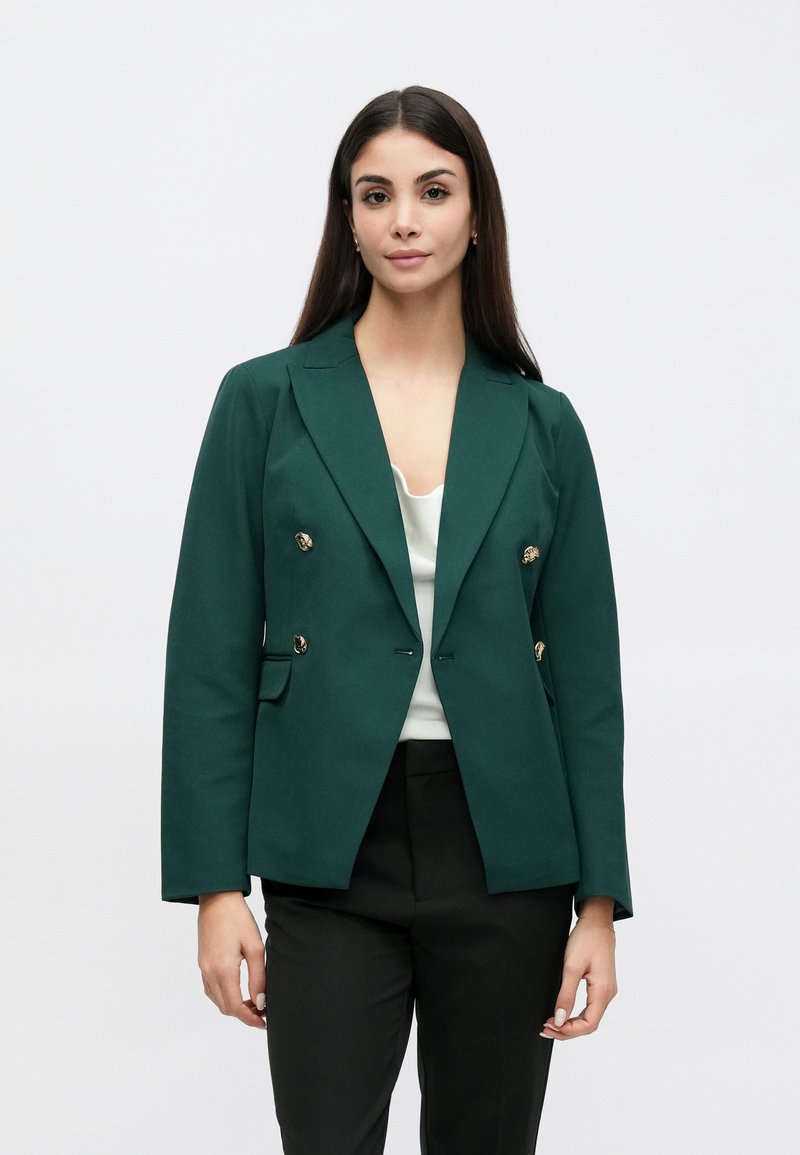 Blazer vert croisé en tissu texturé avec des boutons dorés, des revers crantés et deux poches avant, porté avec un haut clair.