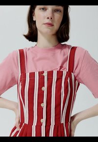 Młoda kobieta o krótkich brązowych włosach nosi różową koszulkę pod czerwono-białą, pionowo paskowaną, bezrękawnikową sukienkę zapinaną na guziki z przodu.