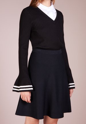 Femme portant un pull noir côtelé à col en V blanc et manches évasées rayées, associé à une jupe noire en ligne A.