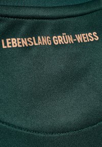 Grön tyg med en slät textur har texten "LEBENSLANG GRÜN-WEISS" i ljusbrunt, placerad på den övre delen av plagget.