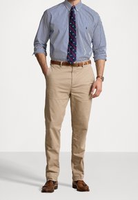 Man som bär en blå och vit randig skjorta med knappar, en marinblå mönstrad slips, beige chinos, ett brunt läderbälte och bruna loafers med tofsar.