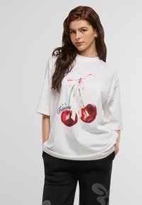 Hvit oversized t-skjorte med et kirsebærillustrasjon med en rosa sløyfe, kombinert med svarte joggebukser som har et subtilt mønster.