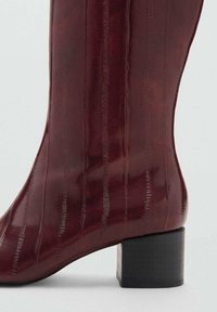 Burgundy-nahka ankle boot, jossa on kiiltävä pinta, uritettu tekstuuri ja paksu musta korko. Sivuprofiilissa korostuu kaareva muoto.