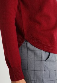 Czerwony dziany sweter z fakturowanym brzegiem i ściągaczami przy rękawach, zestawiony z szarymi spodnimi w kratę o wyraźnym geometrycznym wzorze.