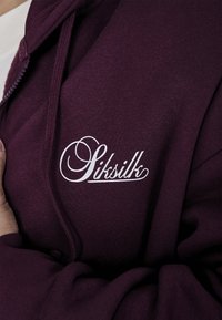 Burgundski zip-over dukserica s bijelim izvezenim logom "Siksilk" na prsima. Mekana tkanina s teksturiranim završetkom i rebrastim manžetama.