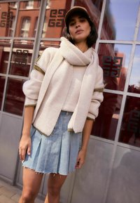 Strickpullover in Creme mit goldenen Akzenten, kombiniert mit einem hellblauen, plissierten Jeansrock. Das Model trägt einen passenden Schal und eine hellbraune Mütze.