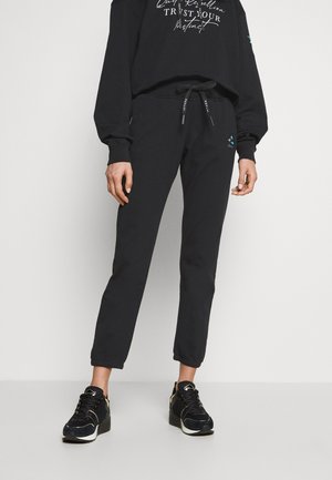 Persoon die zwarte joggerbroek met trekkoord en zwarte sneakers draagt, staand tegen een effen witte achtergrond.