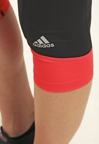 Nærbillede af en person iført sorte og røde Adidas træningsshorts, hvor Adidas-logoet vises på det sorte stof nær knæet.