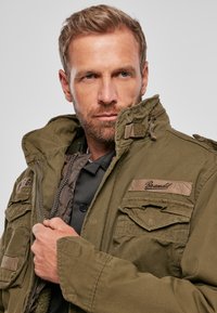 Olive green military-style jacket, featuring multiple front pockets, zip closure, and textured fabric with brand label on the chest.