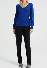 Blå metallisk stickad långärmad tröja med V-hals, kombinerad med svarta byxor med sidoklipp och silvriga spetsiga ankelstövlar.