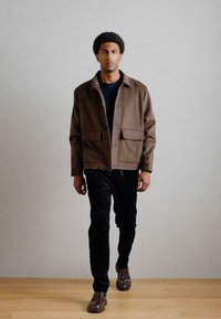 Casaco castanho de botões com dois bolsos frontais, usado sobre uma camisa escura e calças pretas, combinado com sapatos castanhos sobre um fundo de chão de madeira.