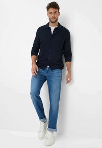 Marineblauwe knoopjesoverhemd met lange mouwen, gecombineerd met lichtblauwe jeans. Model draagt witte sneakers, met opgerolde pijpen van de jeans.