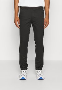 Pantalons noirs ajustés avec une texture lisse, coupe slim, poches latérales et une taille avec passants de ceinture, associés à des chaussures de sport blanches et bleues.