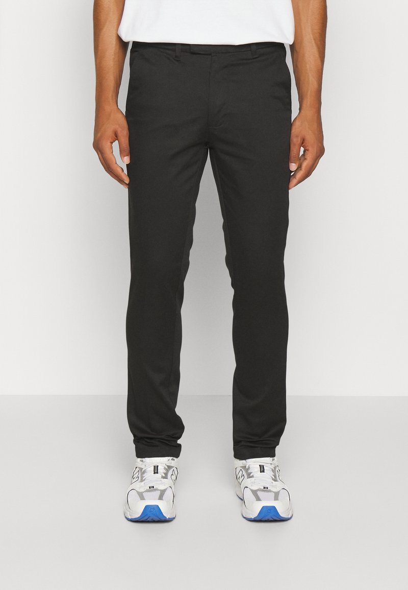 Pantalons noirs ajustés avec une texture lisse, coupe slim, poches latérales et une taille avec passants de ceinture, associés à des chaussures de sport blanches et bleues.