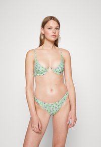 Bikini floral verde com alças ajustáveis; dispõe de um top triangular e uma parte de baixo de cintura baixa. O tecido tem uma textura suave e lisa com um padrão xadrez.