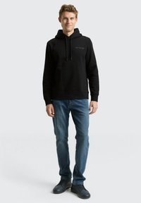 Schwarzer Kapuzenpullover mit Logo auf der Vorderseite, kombiniert mit blauen Jeans und schwarzen Turnschuhen. Der Stoff wirkt weich, und der Schnitt ist lässig und bequem.