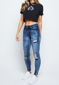 Modré džíny skinny s opotřebovaným vzhledem a několika výřezy, doplněné černým zkráceným tričkem s stříbrným grafickým designem. Bílé tenisky.