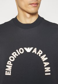 Schwarzes Baumwoll-T-Shirt mit Rundhalsausschnitt. Verfügt über ein cremefarbenes, besticktes Logo, das bogenförmig über der Brust platziert ist. Glatte Oberfläche.