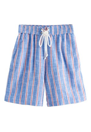 Ung kvinde iført en brun bikini top og blåstribede shorts står på en sandstrand med havbølger og klipper i baggrunden.