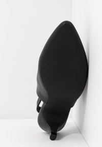Chaussure noire à bout pointu et talon kitten, vue de la semelle, positionnée verticalement contre des surfaces blanches en angle.