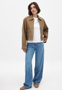 Giacca corta in suede marrone con colletto a punta, camicia bianca sotto, jeans in denim blu larghi e calzature beige. Dettagli di cucitura semplici visibili.