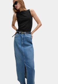 Vrouw in een zwarte mouwloze top, lange blauwe denimschoot met een split aan de voorkant, zwarte riem en zonnebril tegen een effen lichte achtergrond.