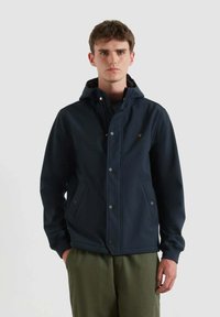 Uomo in giacca con cappuccio blu navy con bottoni a pressione, tasche laterali e pantaloni verde oliva, in piedi di fronte a uno sfondo chiaro e semplice.