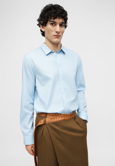 Jeune homme aux cheveux bouclés portant une chemise bleu clair boutonnée, rentrée dans un pantalon marron taille haute avec une ceinture en cuir rembourré.