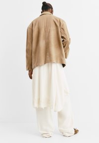 Giacca in suede color sabbia con una vestibilità comoda, indossata sopra un vestito bianco fluido con bordi in pizzo, abbinata a pantaloni ampi color crema e scarpe marroni.