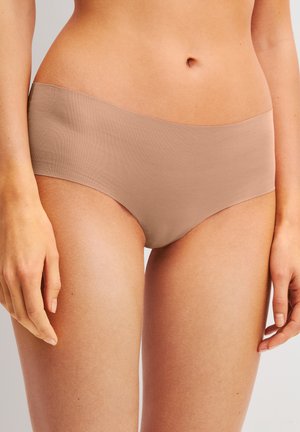 Femme portant un sous-vêtement hipster sans couture couleur chair, debout les mains détendues le long du corps.