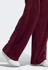 Pantalon large bordeaux avec panneaux latéraux transparents, finition texturée et chaussures de sport blanches. Le pantalon présente des rayures le long des bords latéraux.
