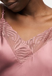 Posnetek ženske v roza majici s tankimi nastavljivimi naramnicami in detajlno čipko z motivom listov vzdolž izreza na vratu.
