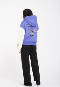 Bluza z kapturem w purpurowym kolorze z białymi rękawami, z graficznymi wzorami na plecach. W zestawie z czarnymi spodniami typu wide-leg i czarnymi butami.