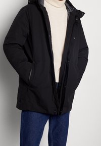 Chaqueta negra impermeable con capucha, cierre de cremallera y bolsillos laterales. Presenta un suave forro interior y un corte holgado, llevada sobre un suéter color crema.