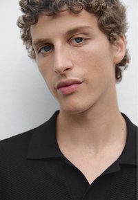Primo piano di una persona che indossa una polo nera con texture e colletto. Carnagione chiara, capelli corti e ricci, e sfondo neutro.