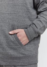 Sudadera gris con tejido texturizado, que presenta un bolsillo lateral y puños acanalados. Diseño simple con detalles de marca mínimos.