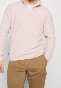 Pullover in pile rosa chiaro con zip a un quarto. Presenta un piccolo logo bianco sul lato sinistro del petto, una texture liscia e maniche lunghe.