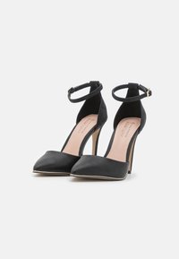 Ein Paar schwarze High-Heels mit spitzen Zehen und Knöchelriemen, auf einem weißen Hintergrund präsentiert.