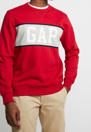 Mand iført en rød sweatshirt med et gråt og sort panel og et hvidt "GAP"-logo, kombineret med beige bukser, står med hænderne i lommerne.