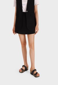 Blazer senza maniche nero su una camicia chiara, abbinato a una minigonna nera e sandali con plateau aperti e dettagli e cinghie marroni.