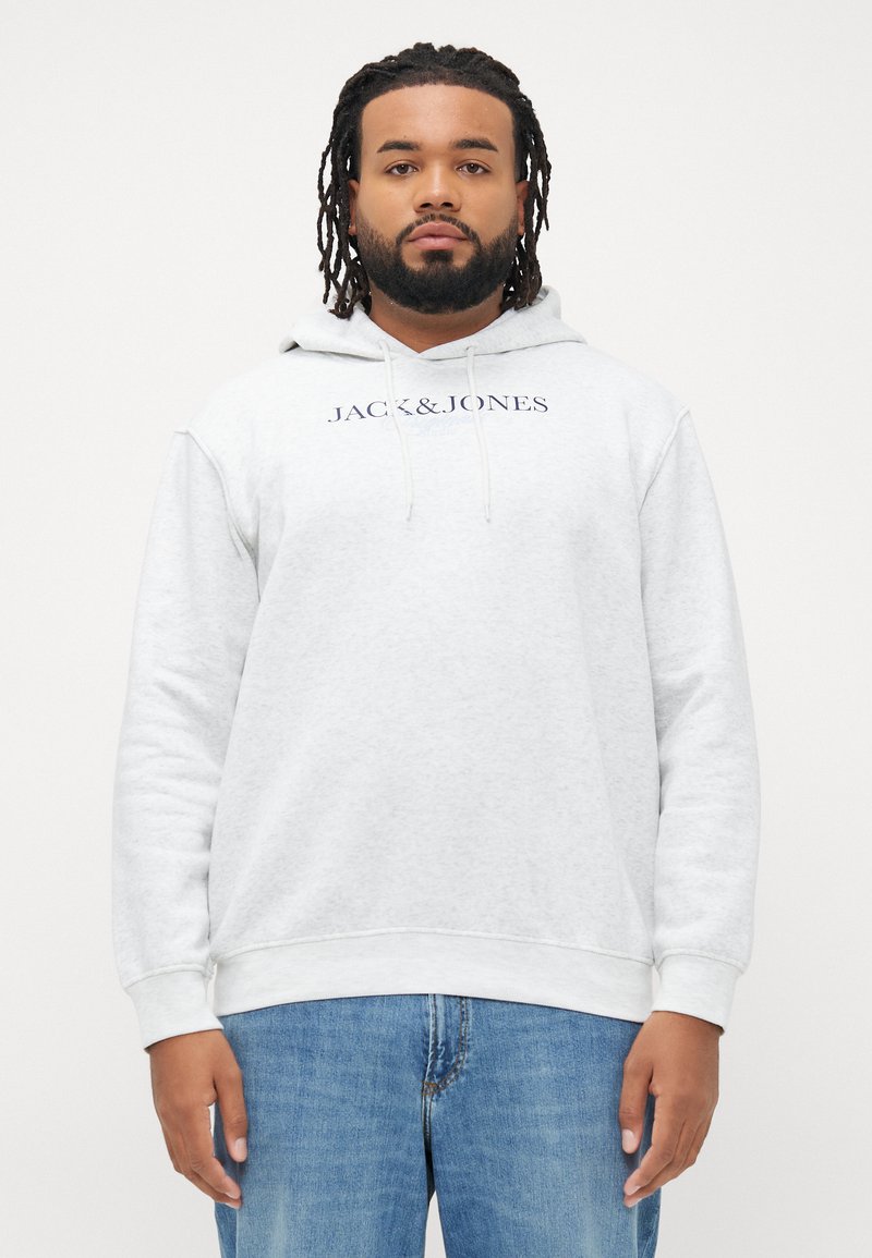 Mann som har på seg en lysegrå hettegenser med "JACK&JONES" tekst og blå jeans, står mot en ensfarget hvit bakgrunn.
