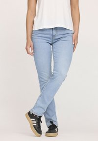 Personne portant un jean bleu clair, des baskets noires avec des rayures blanches, se tenant les jambes croisées, vêtue d'un haut blanc.