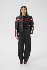 Woman wearing a black leather jacket with red and white stripes, wide black pants, burgundy top, and burgundy pointed shoes standing facing forward.