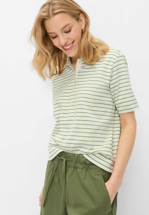 Kvinde smilende, iført grøn- og hvidstribet kortærmet poloshirt og olivengrønne bukser med snøre, hånd i lomme mod hvid baggrund.