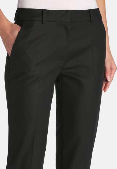 Woman wearing black tailored straight-leg trousers with belt loops, one hand in pocket, shown from waist to mid-thigh.